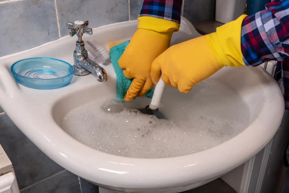 A person with gloves attempting to unblock a sink using a plunger